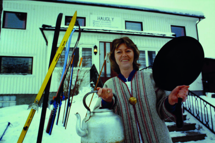 Gammelt bilde fra Hugly Gjesteheim fra 1980-tallet. Vertinne Birgit Lidtveit står oppstilt foran huset med stekepanne og kaffekjele i hendene.