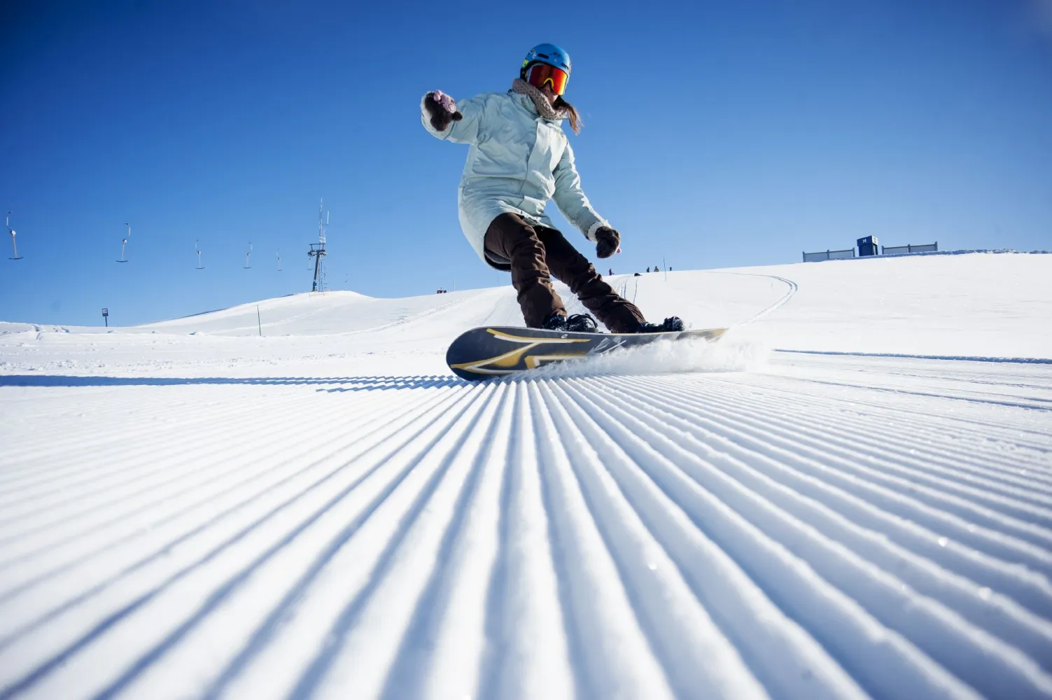 Snowboardkjører i lys jakke som svinger på en nypreparert skiløype under blå himmel.