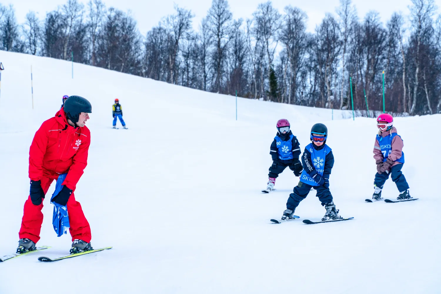 Skiinstruktør i rød skidress viser tre barn hvordan de skal svinge og bremse under skiskole hos Hovden Aktiv.